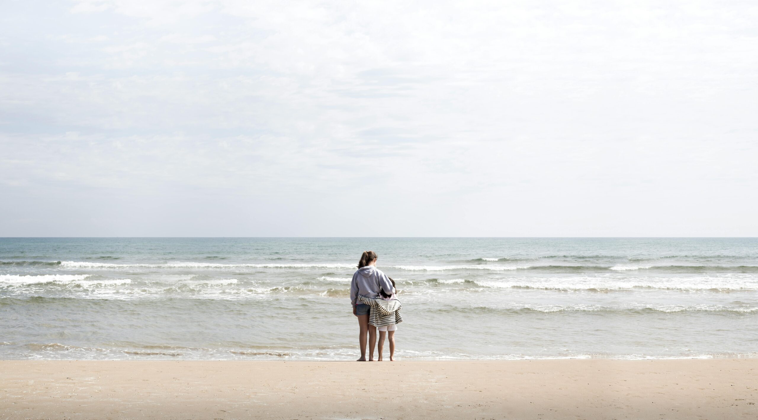 Home A mother and child stand at the shoreline of a Valencia beach, gazing at the waves.